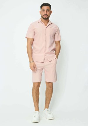 Chemise à boutons à manches courtes rose clair avec une poche poitrine, accompagnée de shorts assortis. Portée avec des baskets blanches, présentant un tissu texturé.