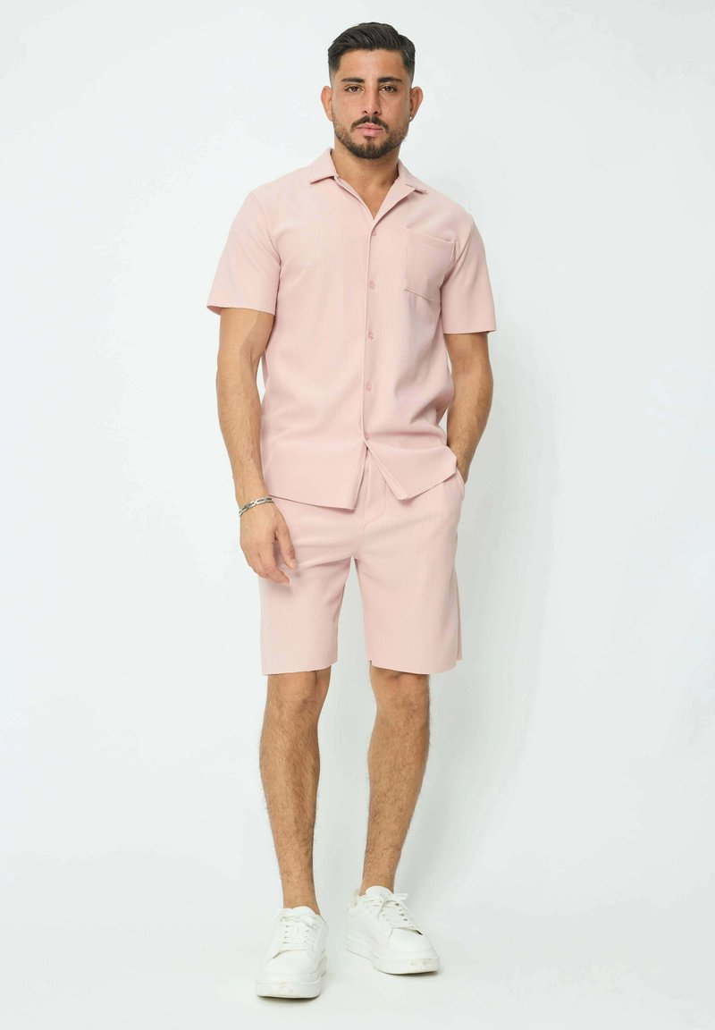 Chemise à boutons à manches courtes rose clair avec une poche poitrine, accompagnée de shorts assortis. Portée avec des baskets blanches, présentant un tissu texturé.