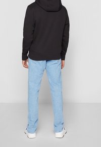 Sudadera negra con bolsillo canguro y cordón ajustable, combinada con jeans de mezclilla azul claro y zapatillas blancas. Se muestra la vista trasera.
