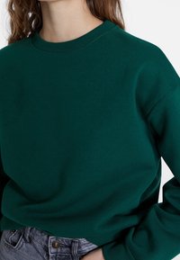 Zelená bavlněná mikina s kulatým výstřihem a žebrovanými manžetami, s volnějším střihem a měkkou texturou. Nasažená přes světle šedé džíny.