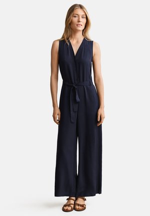 Donna con tuta smanicata blu navy con cintura annodata in vita e sandali marroni con cinturini, in piedi davanti a uno sfondo semplice.