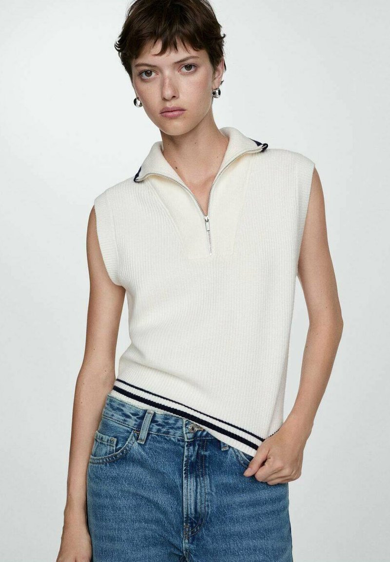 Ærmeløs hvid strikpullover med lynlås krave, der har en ribbet tekstur og marineblå stribe accenter ved kanten. Bæret med blå jeans.