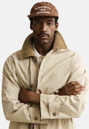 Mann mit Schnurrbart, der einen beigen Mantel mit hochgestelltem Kragen und eine braune Mütze trägt, auf der „Gant International Style 1949 American Sportswear“ steht, mit verschränkten Armen.