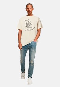 Cremefarbenes Kurzarm-T-Shirt mit einem Hund und einem Surfbrett-Design, kombiniert mit hellblauen, abgewetzten Jeans und weißen Sneakers.