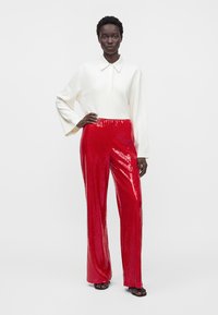 Donna in piedi con la mano sul fianco, indossa un top bianco a maniche lunghe con colletto e pantaloni larghi rossi lucidi con sandali neri.