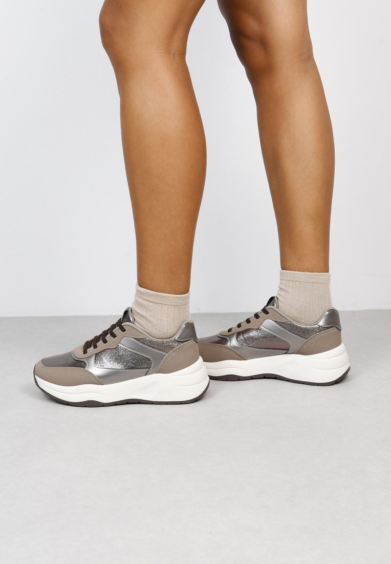 Zapatillas deportivas de cordones en gris y plateado metálico, con una suela blanca gruesa, paneles de malla y texturas contrastantes. Los cordones marrones completan el diseño.