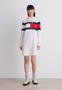 Abito felpa bianco con strisce rosse, blu e nere, caratterizzato dal testo 'TOMMY JEANS'. Abbinato a stivali bianchi con lacci spessi.