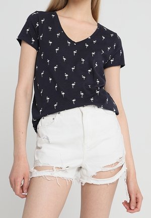 Vrouw draagt een marineblauw T-shirt met V-hals en kleine witte flamingo prints, gecombineerd met versleten witte spijkershorts.