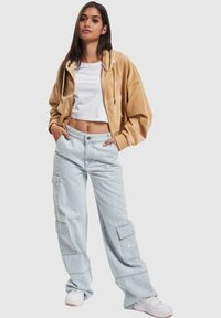 Haut en velours côtelé beige croppé avec une fermeture éclair à l'avant, associé à un pantalon cargo bleu clair avec des poches latérales et un T-shirt blanc en dessous.