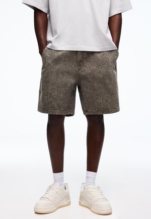 Mand iført gråvaskede knælange shorts, hvide crew-sokker, hvide sneakers og en løs hvid T-shirt med hænderne i lommerne.