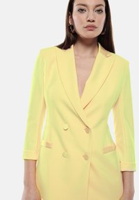 Blazer doppiopetto giallo chiaro con revers a punta, maniche tre quarti, due tasche frontali e bottoni tondi. Tessuto liscio.