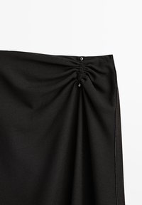 Tissu noir avec détail froncé maintenu par deux petits boutons noirs près du bord, montrant la texture et les coutures sur un fond blanc.