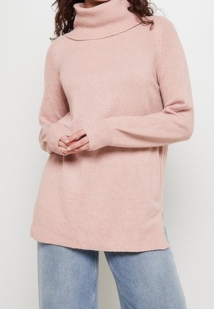 Žena v jemně růžovém roláku a světle modrých džínách s rukama jemně sepnutýma před sebou.