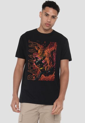 Young man wearing a black Batman graphic t-shirt with fiery orange and red design, paired with beige cargo shorts, standing against plain background.