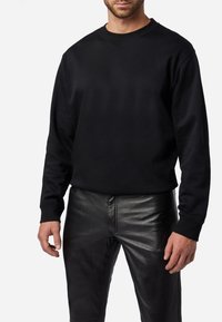 Schwarzes Sweatshirt mit rundem Halsausschnitt und langen Ärmeln, aus weichem Stoff gefertigt. Dazu passend glänzende, enganliegende schwarze Lederhosen.