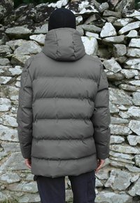 Jachetă puffer gri cu secțiuni quilted orizontale și glugă, purtată peste pantaloni negri, pe un fundal de zid din piatră.