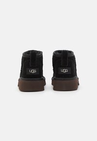 Botas de tobillo de ante negro con acabado texturizado, tacón reforzado y suela de goma marrón. Presenta el logo de UGG en la parte trasera.