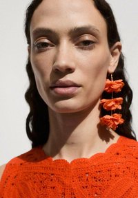 Luftiges gehäkeltes Oberteil in leuchtendem Orange mit einem strukturierten floralen Muster; hängende Ohrringe aus geschichteten orangen Stoffblättern.