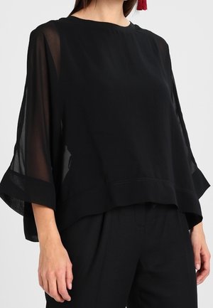 Femme portant un chemisier noir ample avec des manches larges transparentes et un pantalon noir, debout devant un fond clair uni.