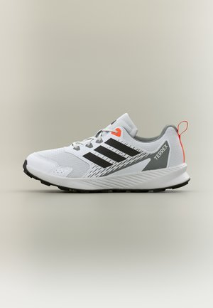 Λευκό και γκρι αθλητικό παπούτσι Adidas Terrex με μαύρες γραμμές, πορτοκαλί οφθαλμό κορδονιού και ταινία έλξης, εμφανιζόμενο σε πλευρικό προφίλ σε ουδέτερο φόντο.