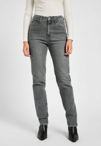 Vaqueros negros de cintura alta en un lavado desvanecido, diseño de pierna recta, con cinco bolsillos y costuras visibles. Llevados con una blusa ajustada y botas.