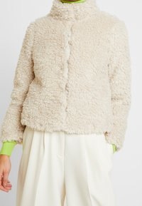 Beige fuskpälsjacka med ståkrage, tryckknappsstängning och långa ärmar som har gröna stickade detaljer vid manschetterna. Mjuk, texturerad yta.