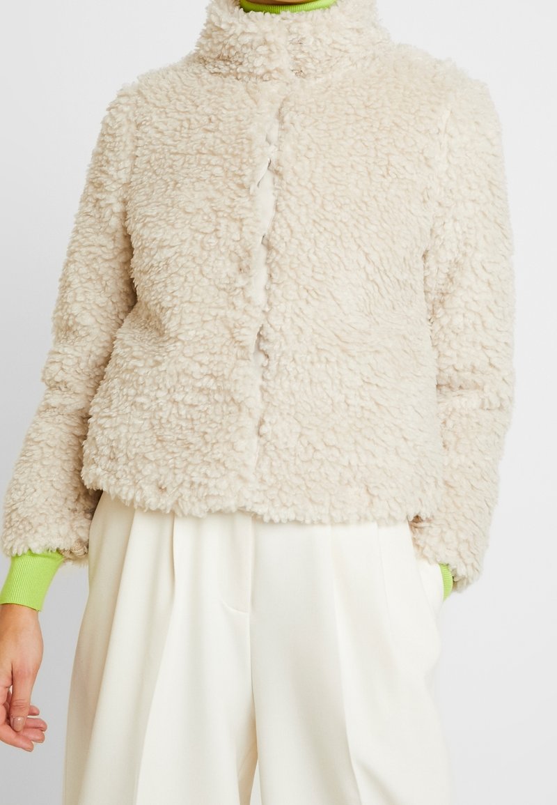 Beige fuskpälsjacka med ståkrage, tryckknappsstängning och långa ärmar som har gröna stickade detaljer vid manschetterna. Mjuk, texturerad yta.