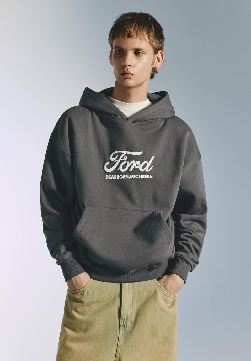 Jeune personne portant un sweat à capuche gris Ford avec le texte « Dearborn, Michigan » et un pantalon beige clair, les mains dans les poches, devant un fond uni.