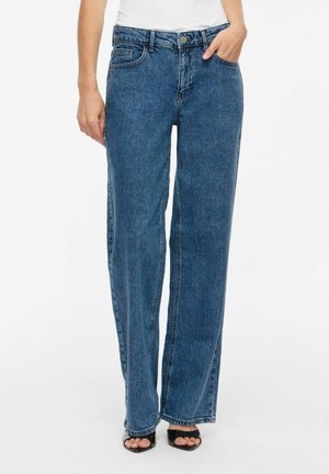 Femme portant un jean bleu taille haute à jambes droites, un haut blanc et des talons noirs ouverts à l’avant, une main dans la poche.