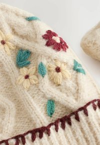 Off-white strikket stof med farverig blomsterbroderi i rød, gul og grøn. Har en tekstureret overflade og en bølget maroon kant.