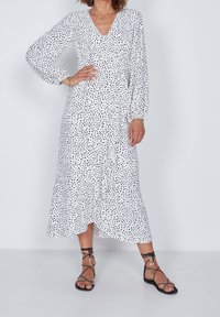 Vestido de lunares en blanco y negro con mangas largas, escote en V y dobladillo fluido. Combinado con sandalias negras de tiras, tono de piel visible.