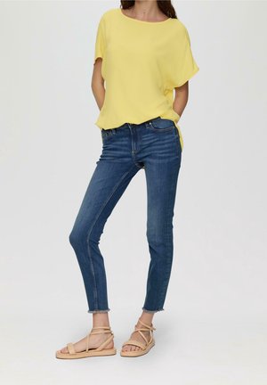 Kobieta ubrana w żółty top z krótkim rękawem, niebieskie obcisłe dżinsy z postrzępionymi nogawkami oraz beżowe, płaskie sandały z paskiem wokół kostki, stoi z rękami w kieszeniach.