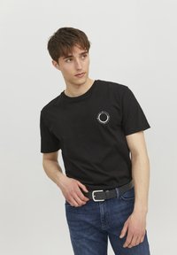 Camiseta de algodón negra de manga corta, con cuello redondo y un pequeño logo circular blanco en el lado izquierdo del pecho. Combinada con unos vaqueros azules y un cinturón negro.
