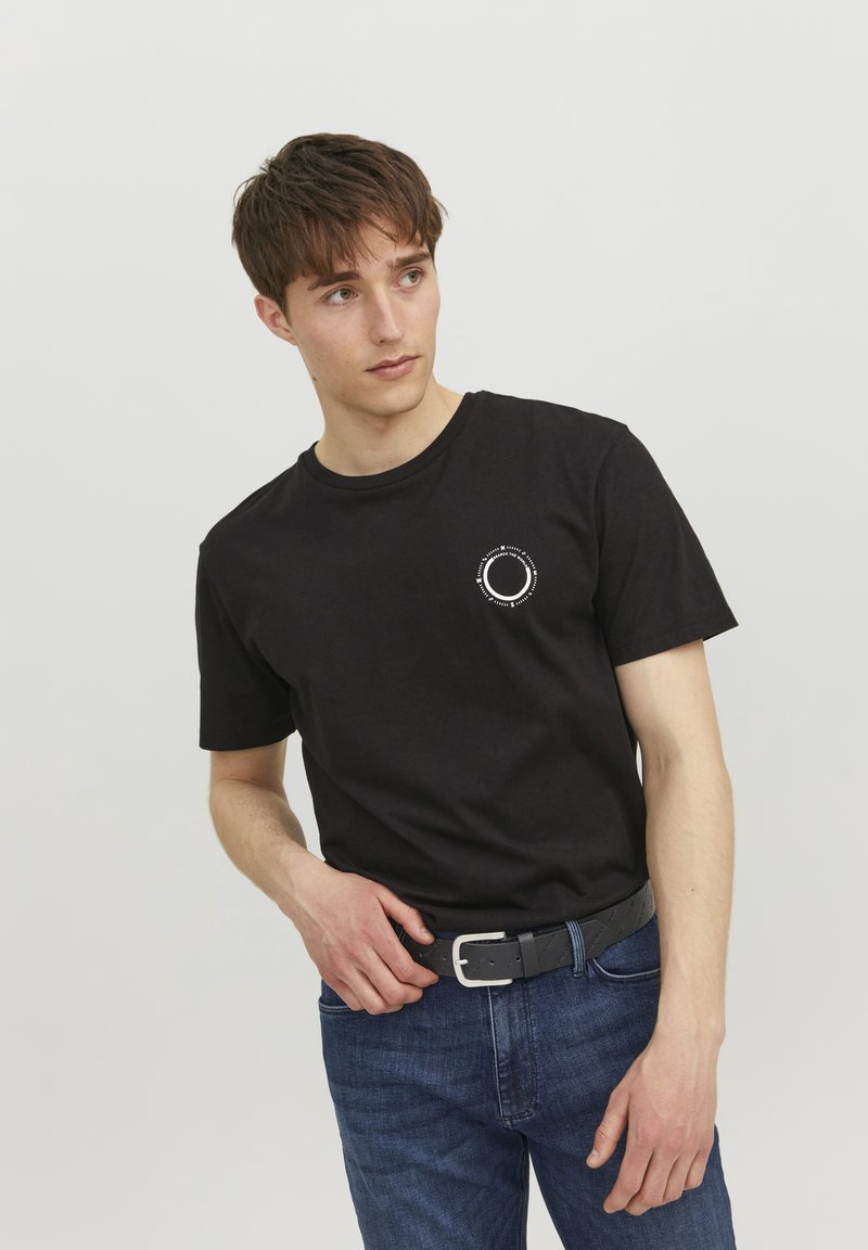 Camiseta de algodón negra de manga corta, con cuello redondo y un pequeño logo circular blanco en el lado izquierdo del pecho. Combinada con unos vaqueros azules y un cinturón negro.