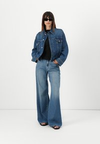 Giacca di denim blu medio con due tasche frontali, abbinata a un top nero a coste e jeans a gamba larga. Sandali neri completano il look.