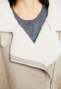 Beige faux suede jacket with a white, fluffy collar, zippered front, and gray sweater underneath, featuring a round knit neckline and button.