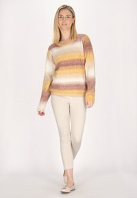 Pulover tricotat cu dungi multicolore în galben, maro și crem. Are un decolteu rotund și mâneci lungi. Este purtat cu pantaloni slim-fit bej.