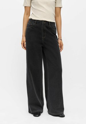 Femme portant un jean denim noir à jambes larges avec des coutures visibles, associé à un haut manches courtes beige clair et des chaussures noires.