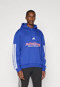 Modra kapuca s puloverjem z belimi črtami na rokavih in velikim rdečim napisom "ADIDAS SPORTSWEAR" na sprednji strani, pletenina.