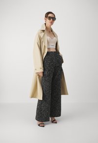 Beige trenchcoat, gewatteerde korsettop en wijdvallende zwart-witte textuurbroek. Zwarte sandalen met bandjes maken de outfit compleet.