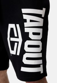 Juodos sportinės šortai su dideliu baltu "TAPOUT" užrašu kairėje pusėje, pagaminti iš minkštos, lygaus audinio.
