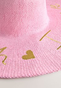 Růžový sluneční klobouk z tkaniny s vyšíváním zlatých srdcí a písmen. Materiál je texturovaný a hustě pletený, s širokou krempou.