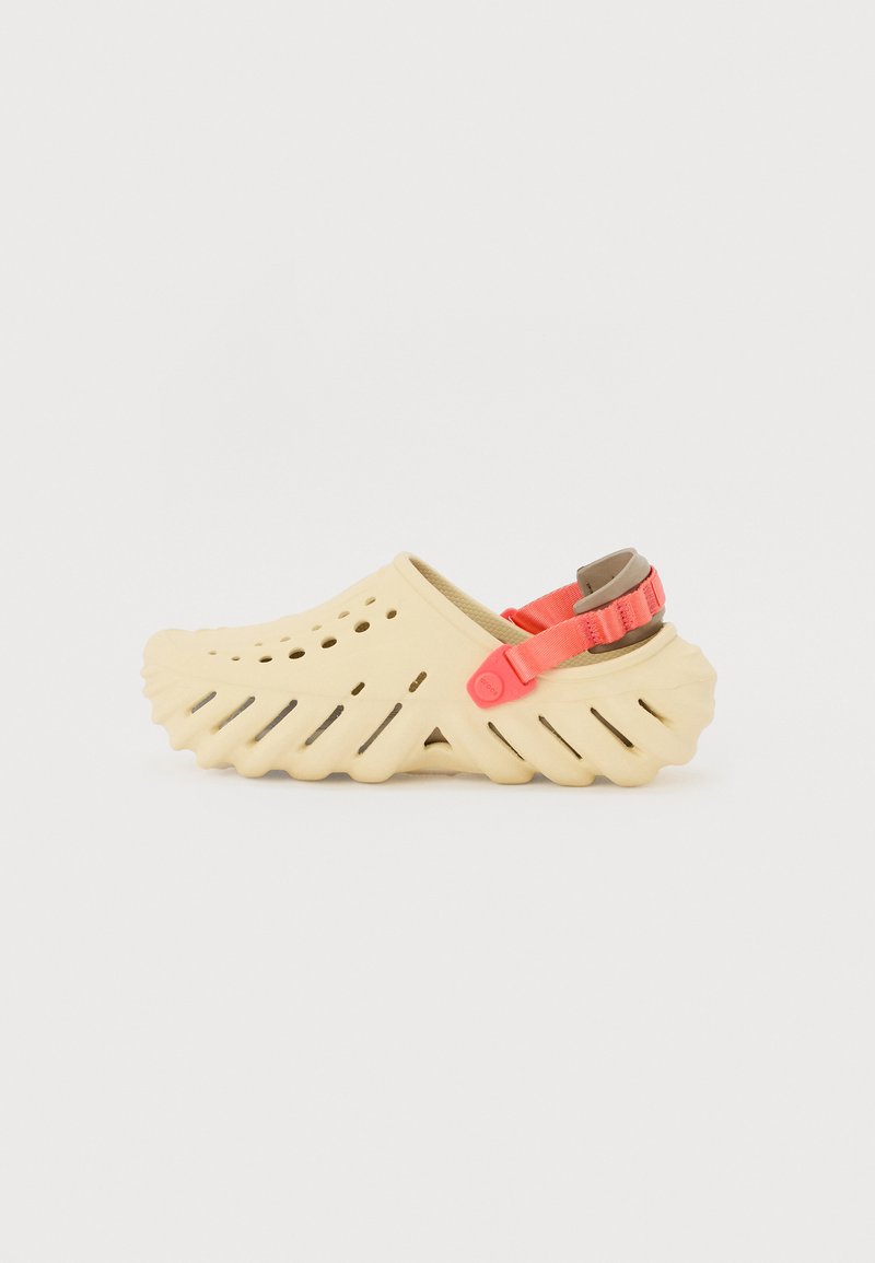 Beige gummitræsko med en bølget sål, der har ventilationshuller, samt et justerbart pink bånd for en sikker pasform.
