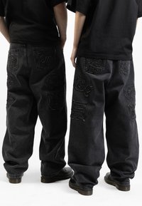 Jeans baggy neri in denim con toppe ricamate e dettagli testurizzati. Presentano un alto punto vita, gambe ampie e tasche posteriori visibili.