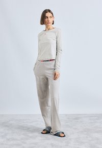 Svijetlosivi dugi rukav majica s logotipom, u kombinaciji sa svijetlosivim slobodnim hlačama s elastičnim pojasom s brandom. Crne klizne cipele.