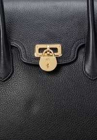 Musta nahkalaukku, jossa on teksturoidun pinnan viimeistely. Varustettu kultaisella padlock-tyylisellä suljennalla ja suorakulmaisilla metalliosilla. Vankat mustat kahvat.