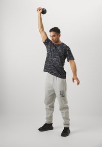 Sort camo fitness t-shirt og grå sweatpants med "UA" logo, båret med sorte sportssko, mens en person løfter en kettlebell over hovedet.