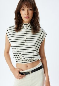Gestreepte crop top in crème en zwart, met een hoge kraag en een gathered zoom. Gepaarde met een witte rok en een zwarte riem met een zilveren gesp.