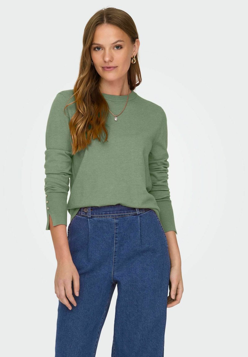 Grön tröja med långa ärmar, ribbade muddar och delad detalj vid handlederna. Matchad med högmidjade denimbyxor.