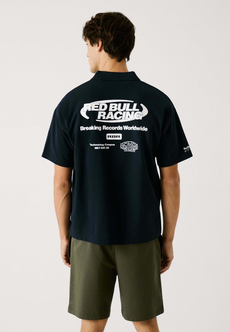 Zwarte shirt met korte mouwen met "RED BULL RACING" in het wit op de achterkant, vergezeld van verschillende logo's en tekst. Draagt in combinatie met olijfgroene shorts.